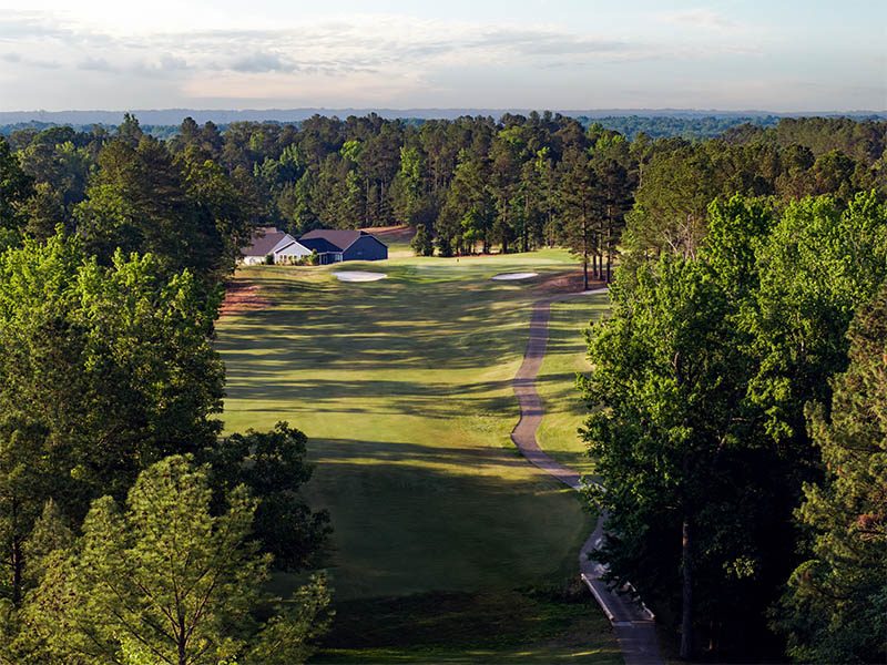 Aerial view of a tree-lined golf fairway with a winding cart path, sand bunkers, and a house in the distance.